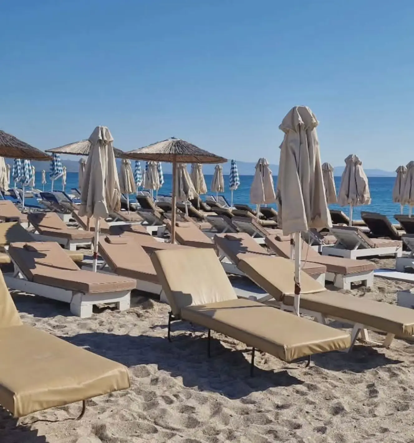 Rows of sunbeds with straw parasols on the sand facing the blue Aegean